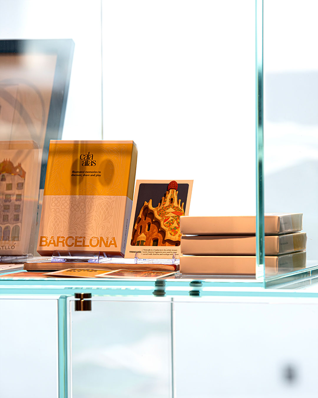 Cala Atlas Barcelona box and Park Güell illustrated card displayed on a glass shelf with stacked boxes and bright background