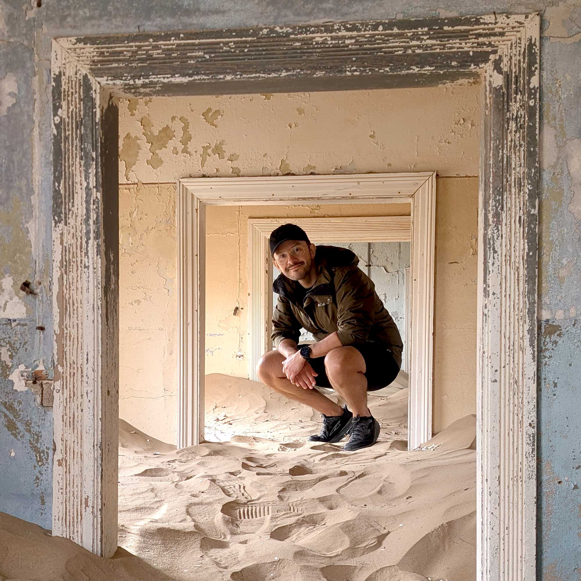 Person crouching inside aKolmanskop building, framed by multiple peeling doorframes