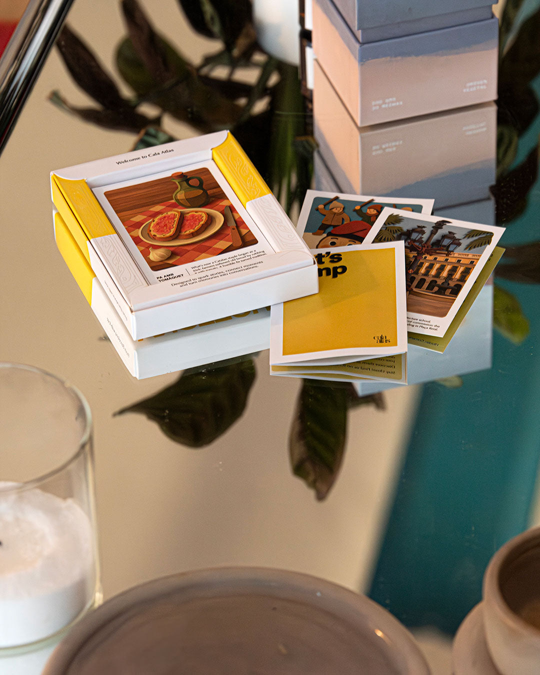 Cala Atlas Barcelona Story Deck box and scattered illustrated cards on a reflective table with plants and ceramics