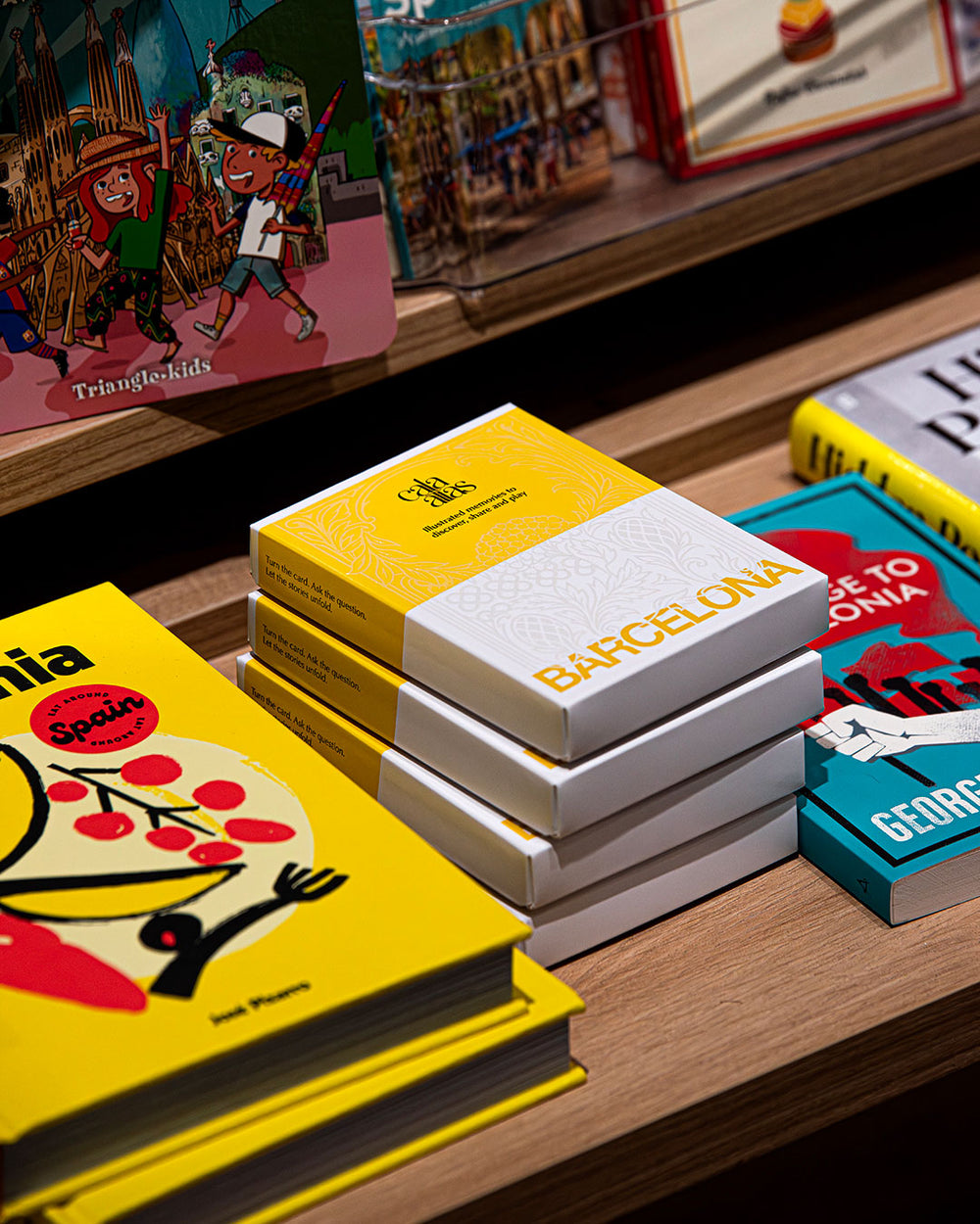 Stacked Cala Atlas Barcelona boxes displayed on a wooden shelf surrounded by colourful illustrated books and covers