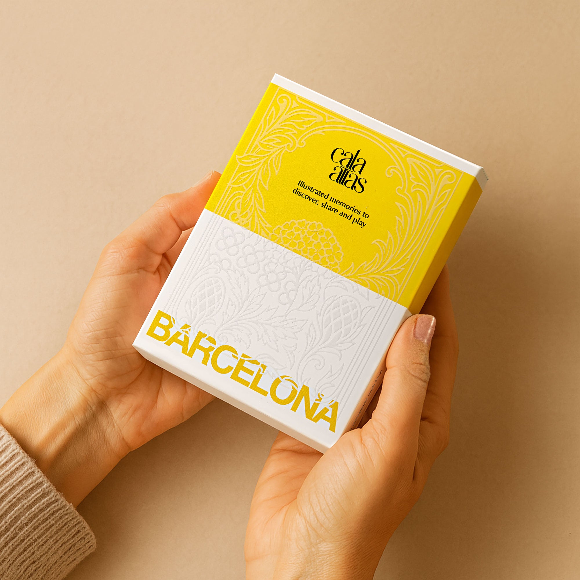 Hands holding the Cala Atlas Barcelona Story Deck gift box, showing the yellow and white embossed packaging