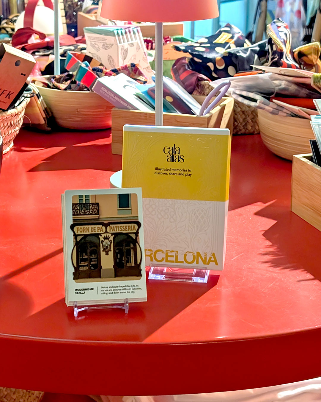 Cala Atlas Barcelona Story Deck and bakery facade illustrated card displayed on a red table surrounded by baskets of accessories