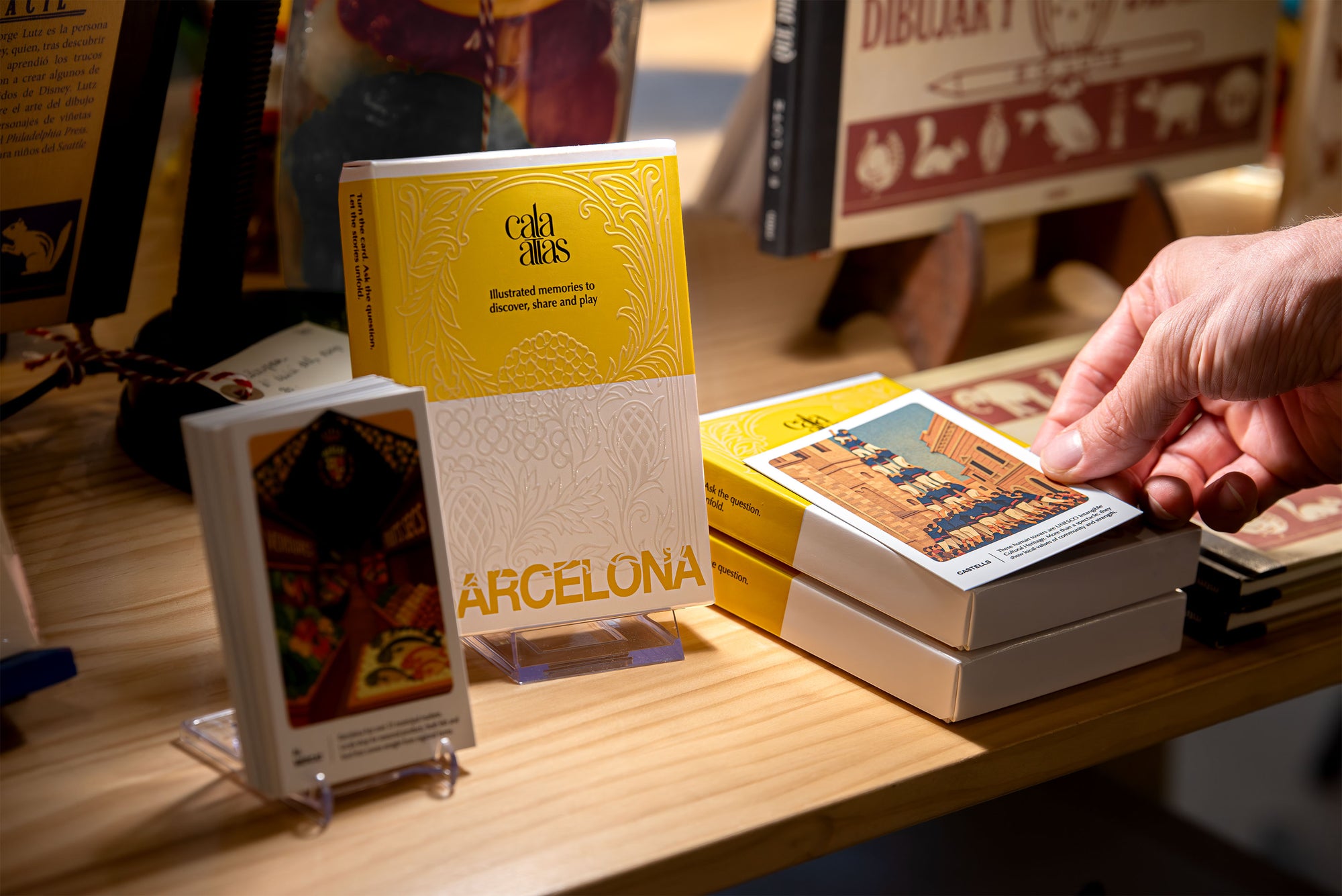 Cala Atlas Barcelona Story Deck displayed on a wooden shop counter with a customer hand picking up an illustrated card
