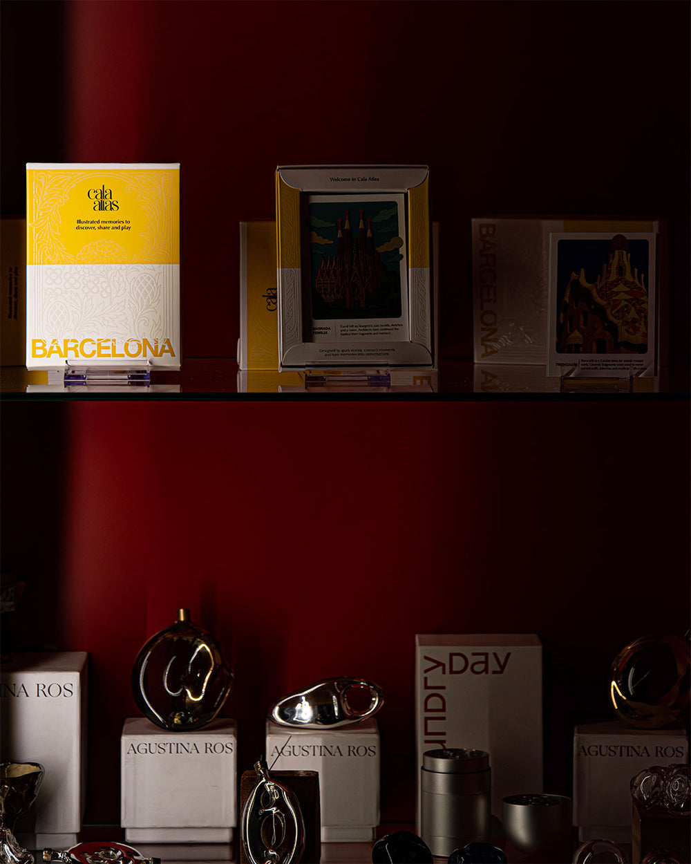 Cala Atlas Barcelona Story Deck and illustrated cards displayed on a dark red shelf beside metallic decorative objects.