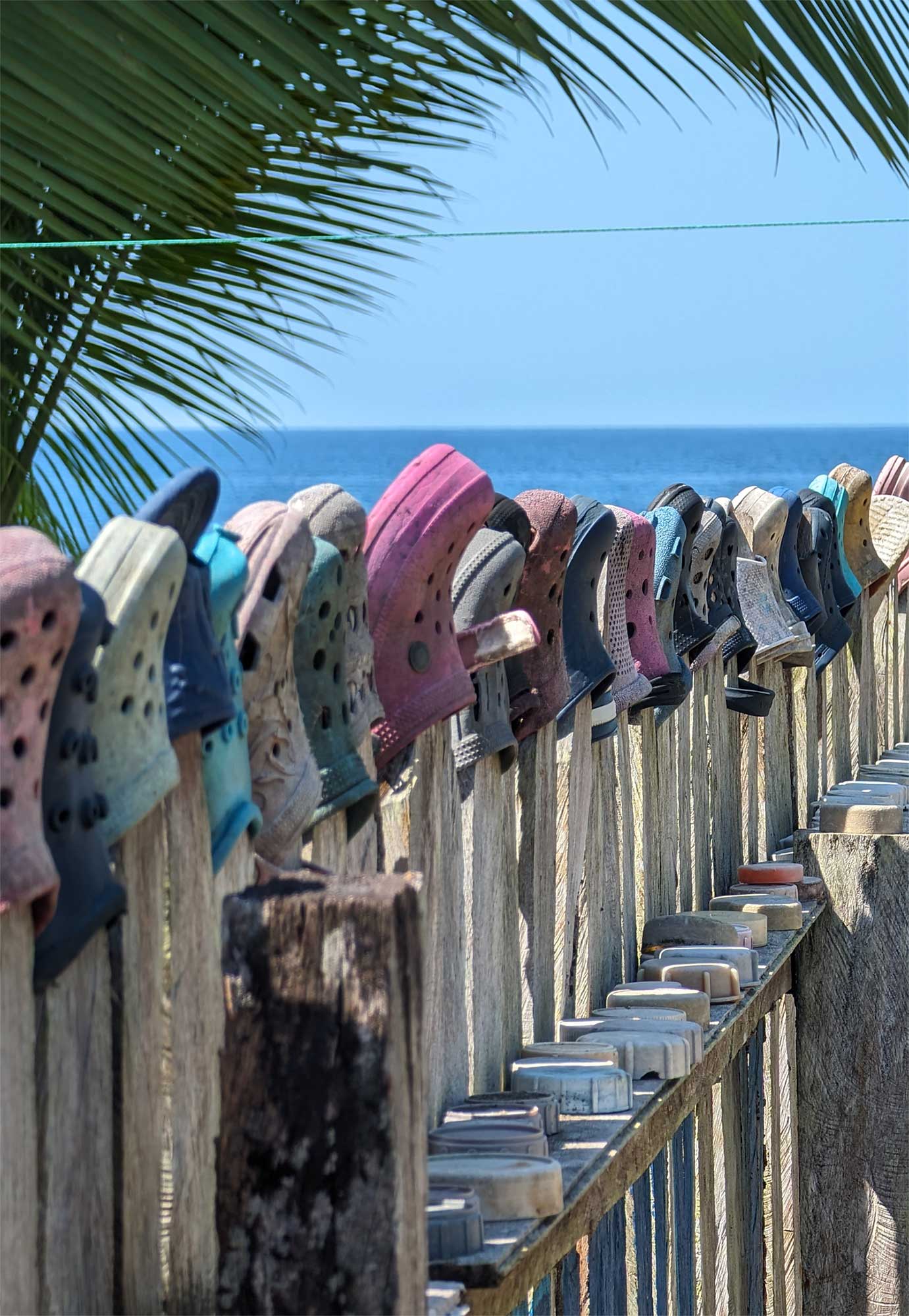 Row of colourful rubber clogs placed along the top of a wooden fence with the ocean visible in the background