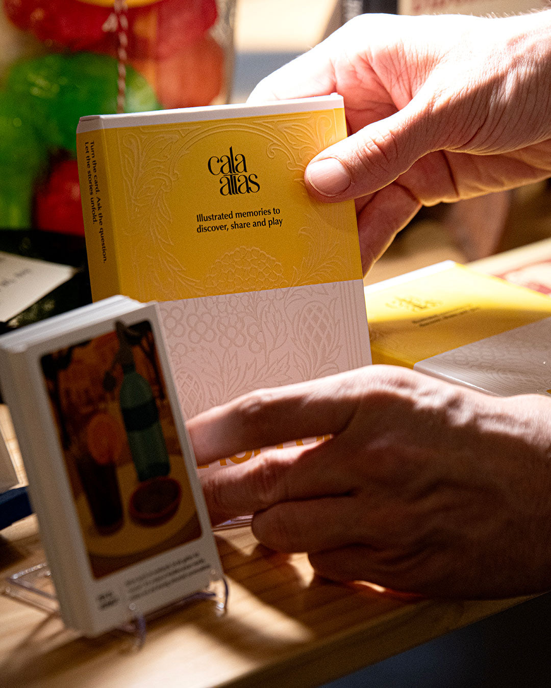 Hands holding a Cala Atlas Barcelona yellow box above a wooden surface with another illustrated card in the foreground.