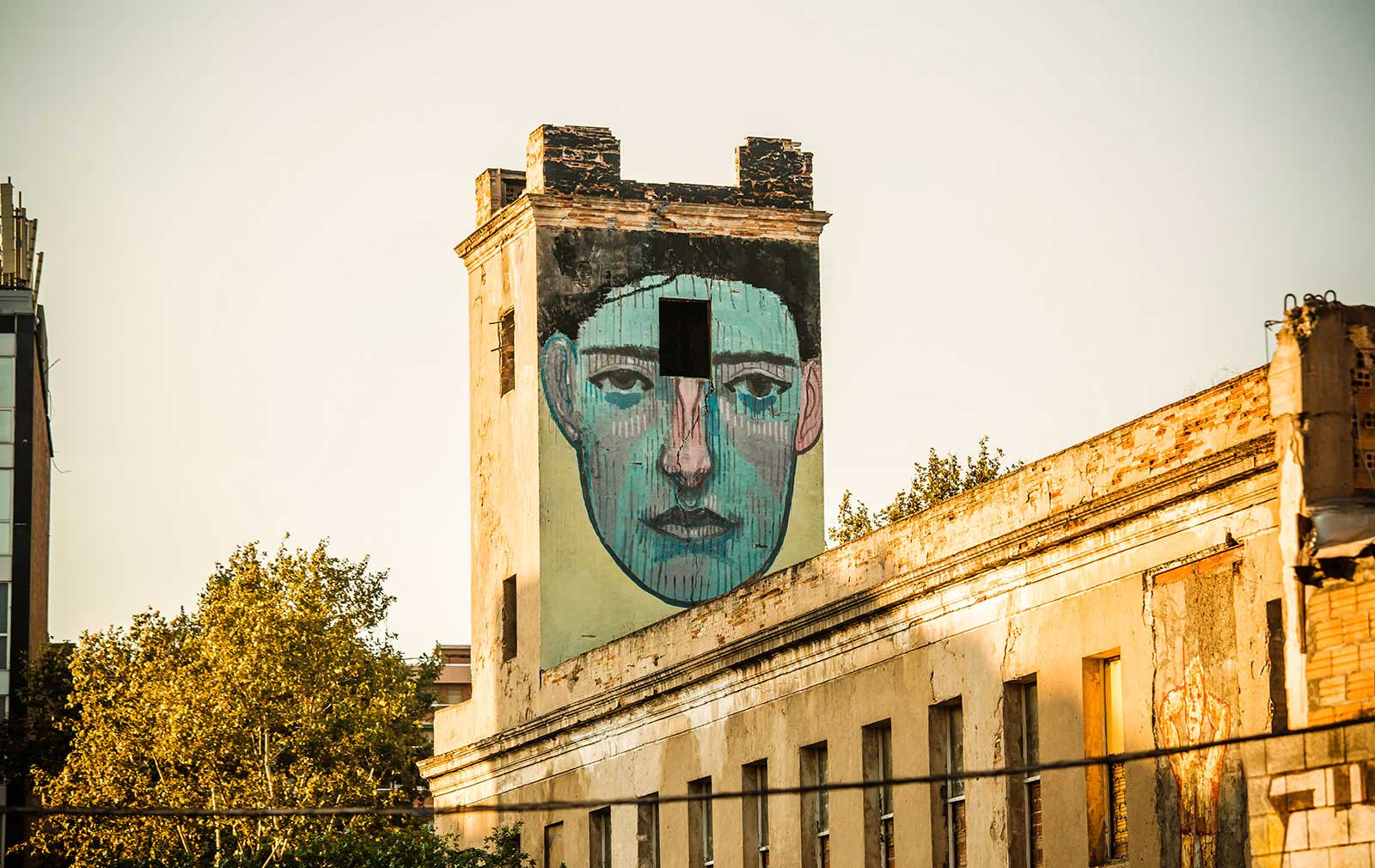 Large painted blue face covering the upper facade of a worn beige building at sunset