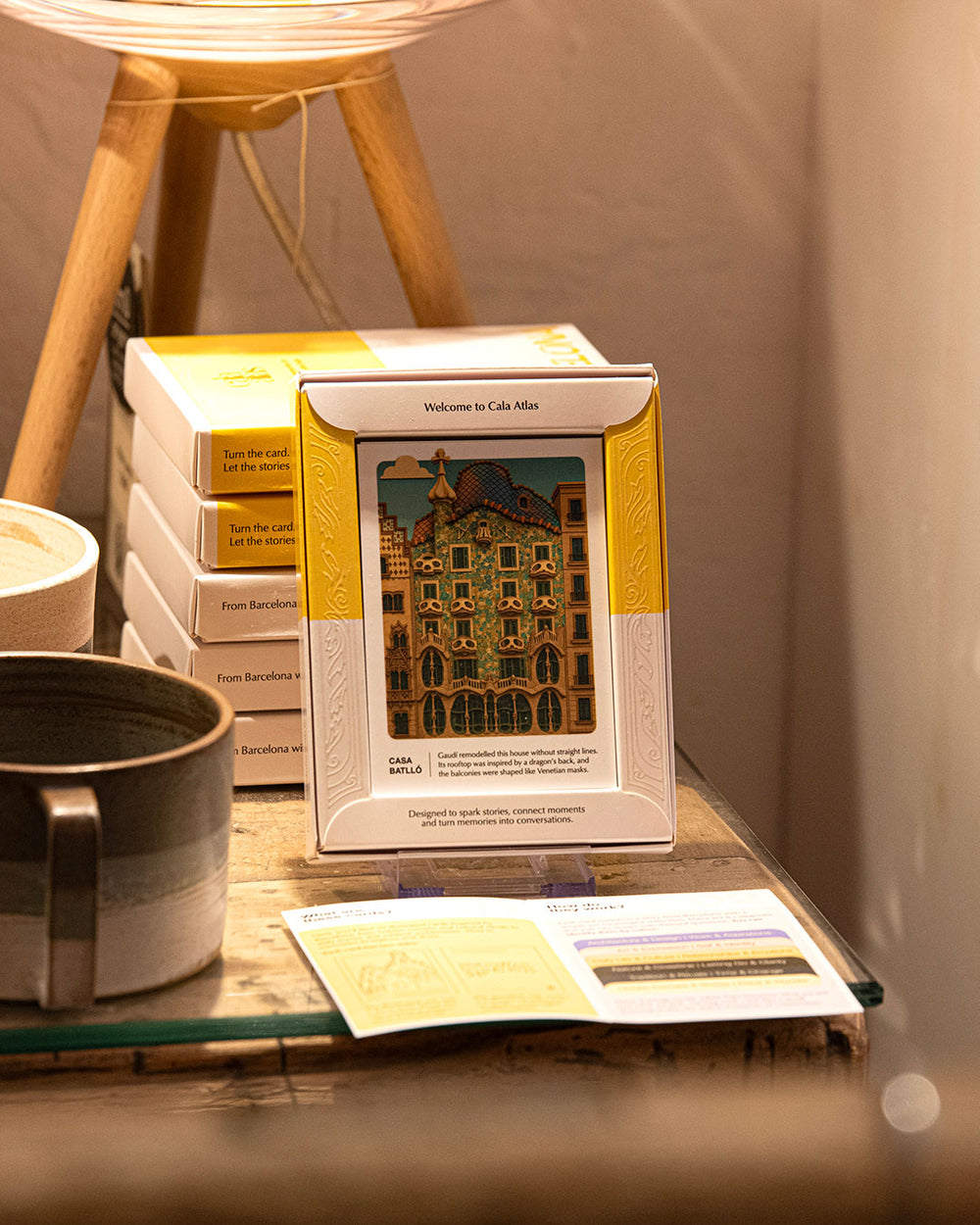 Cala Atlas Barcelona Story Deck box with Casa Batlló illustration beside stacked boxes, ceramic mug and open leaflet