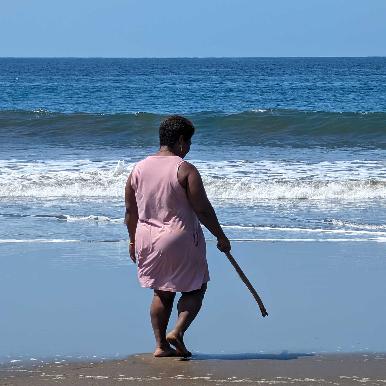 Woman in a pink dress walking barefoot on wet sand toward the shoreline with waves in the background
