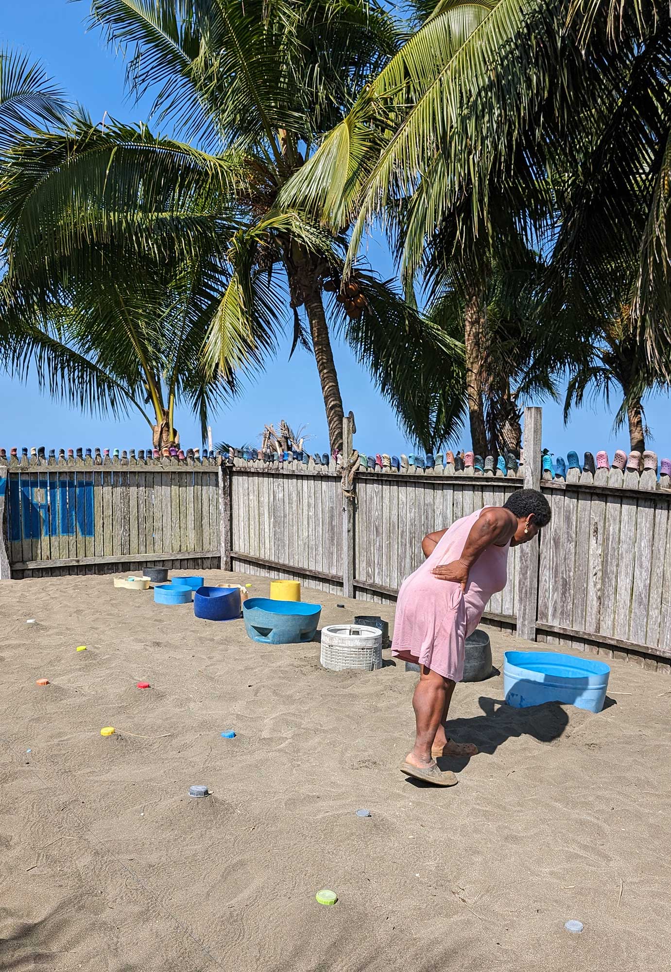 Woman in a pink dress bending forward to inspect coloured sand containers inside a fenced outdoor area with palm trees