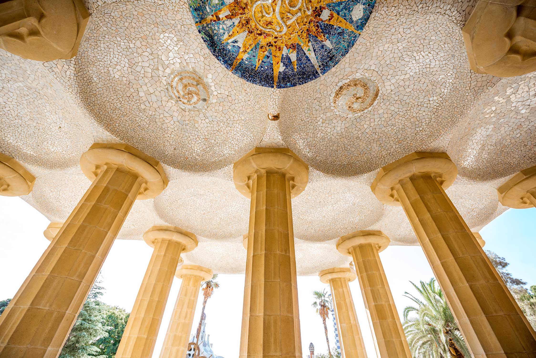 Stone columns supporting a ceiling covered in white mosaic tiles with circular decorative patterns at Park Güell