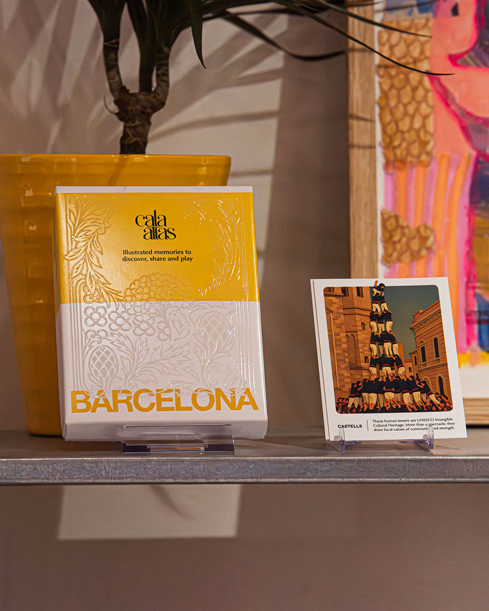 Cala Atlas Barcelona yellow box and illustrated castells human tower card displayed on a shelf with plant pot and artwork behind