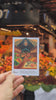Hand holding a Cala Atlas illustrated market card in front of a fresh produce stall inside a market, with fruits and vegetables blurred in the background