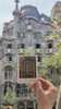 Hand holding a Cala Atlas illustrated Casa Batlló card in front of the real Casa Batlló facade in Barcelona, with the building blurred in the background