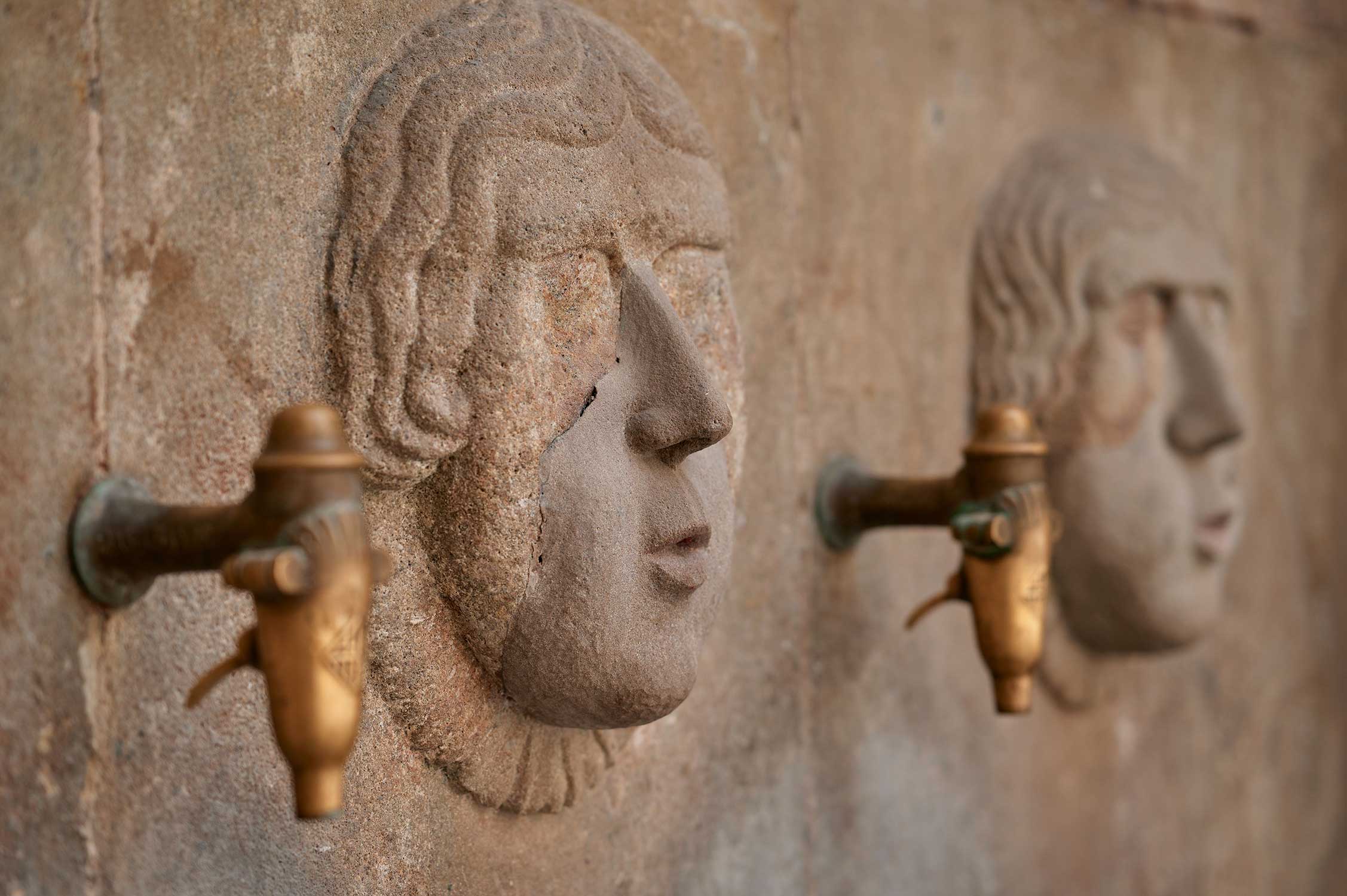 Close-up of a carved stone face with a bronze fountain spout mounted on a textured wall.